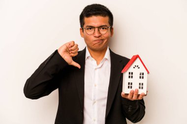 Latin kökenli genç bir adam beyaz arka planda izole edilmiş küçük bir ev tutuyor. Hoş olmayan bir jest yapıyor. Anlaşmazlık konsepti.