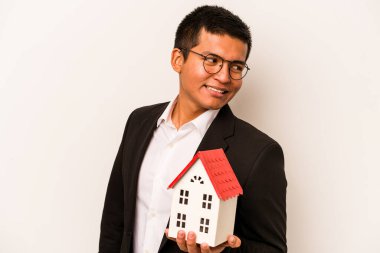 Beyaz arka planda izole edilmiş küçük bir evi tutan İspanyol genç adam gülümsüyor, neşeli ve keyifli görünüyor..