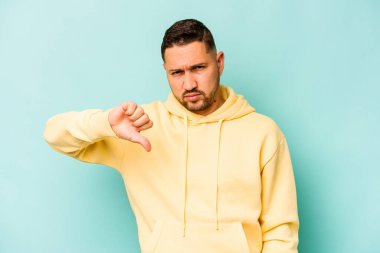 Mavi arka planda izole edilmiş genç İspanyol adam baş parmağını aşağı gösteriyor, hayal kırıklığı konsepti.