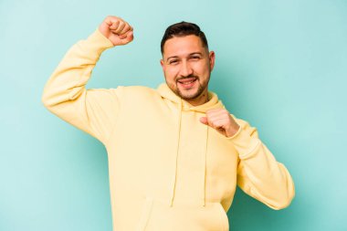 Genç İspanyol adam mavi arka planda izole edilmiş özel bir günü kutluyor, atlıyor ve enerjiyle kollarını kaldırıyor..