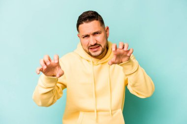 Mavi arka planda izole edilmiş genç İspanyol bir adam pençelerini bir kediyi taklit ederken gösteriyor, agresif bir hareket..