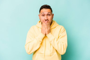 Mavi arka planda izole edilmiş genç İspanyol adam şok olmuş, ağzı ellerle kaplanmış, yeni bir şey keşfetmek için sabırsızlanıyor..