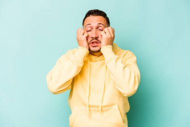 Genç İspanyol bir adam mavi arka planda izole edilmiş, ağlıyor, bir şeyden memnun değil, acı ve kafa karışıklığı konsepti..