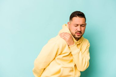 Mavi arka planda izole edilmiş genç İspanyol bir adam omuz ağrısı çekiyor..