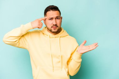 Genç İspanyol bir adam mavi arka planda izole edilmiş ve elinde bir ürün tutuyor..