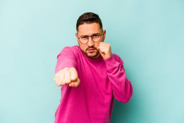 Latin kökenli genç bir adam mavi arka planda izole edilmiş. Yumruk atıyor, sinirleniyor, kavga ediyor..