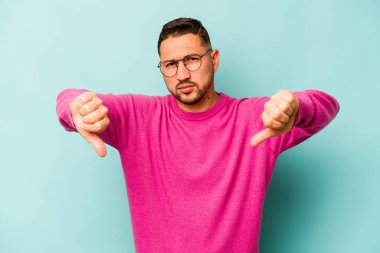 Mavi arka planda izole edilmiş genç İspanyol adam baş parmağını aşağı gösteriyor ve hoşlanmadığını ifade ediyor..