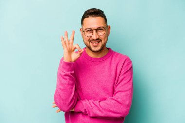 Mavi arka planda izole edilmiş genç İspanyol bir adam bir göz kırptı ve elleriyle iyi bir jest yaptı..