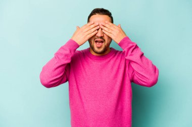 Mavi arka planda izole edilmiş genç İspanyol adam elleriyle gözlerini kapatıyor, geniş bir gülümsemeyle bir sürpriz bekliyor..