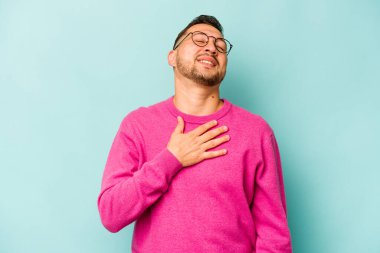Mavi arka planda izole edilmiş genç İspanyol adam yüksek sesle gülüyor ve elini göğsüne koyuyor..