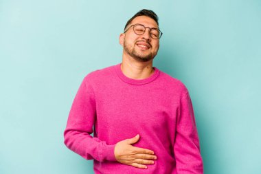 Mavi arka planda izole edilmiş genç İspanyol adam karnına dokunuyor, nazikçe gülümsüyor, yemek yiyor ve tatmin oluyor..