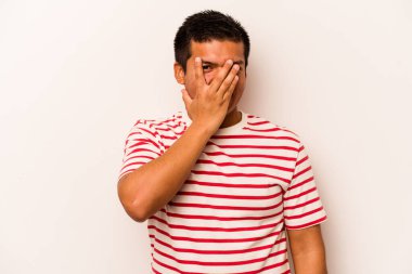Beyaz arka planda izole edilmiş genç İspanyol adam kameraya parmaklarıyla göz kırpıyor, yüzünü kapatmaktan utanıyor..