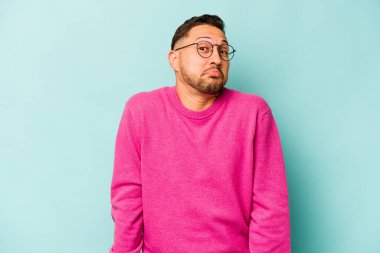 Mavi arka planda izole edilmiş genç bir İspanyol omuz silker ve gözleri açık kalır..