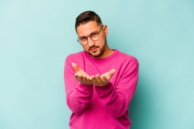 Genç İspanyol bir adam mavi arka planda izole edilmiş. Elinde avuç içi olan bir şeyle kameraya sunuyor..
