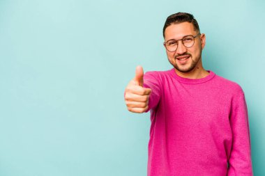 Genç İspanyol adam mavi arka planda izole edilmiş gülümsüyor ve başparmağını kaldırıyor.