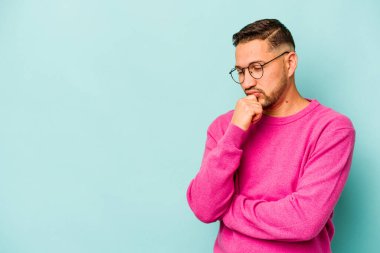 Genç İspanyol adam mavi arka planda izole edilmiş şüpheli ve şüpheci bir ifadeyle yana bakıyor..