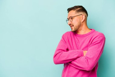 Mavi arka planda izole edilmiş genç İspanyol adam kollarını kavuşturarak gülümsüyor..