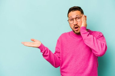 Mavi arka planda izole edilmiş genç İspanyol adam, avuç içinde kopya alanı tutmaktan etkilenmiş..
