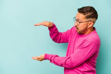 Mavi arka planda izole edilmiş genç İspanyol adam elleri arasında bir kopya alanı tutarken şok oldu ve hayrete düştü..