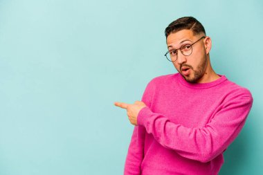Mavi arka planda izole edilmiş genç İspanyol adam gülümsüyor ve boş alanda bir şeyler gösteriyor..