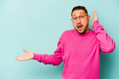 Mavi arka planda izole edilmiş genç İspanyol bir adam avucunun içinde fotokopi alanı tutuyor, el üstünde tutuyor. Şaşırmış ve mutlu..