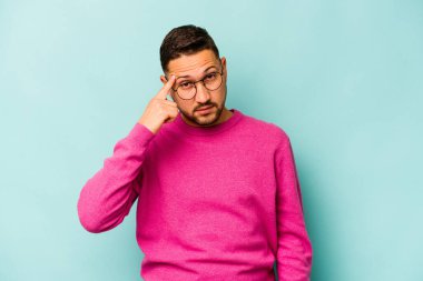 Genç İspanyol bir adam mavi arka planda izole edilmiş, parmağıyla tapınağı işaret ediyor, düşünüyor ve bir göreve odaklanıyor..