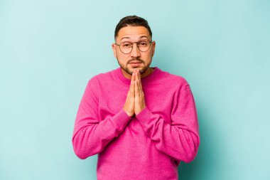 Mavi arka planda izole edilmiş genç bir İspanyol. El ele tutuşup, ağzının kenarında dua ediyor. Kendine güveniyor..