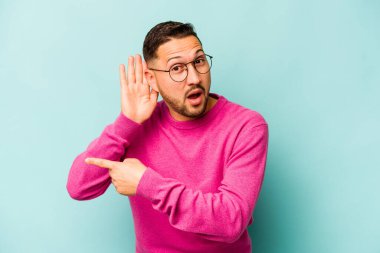 Genç İspanyol bir adam mavi arka planda izole edilmiş bir şekilde bir dedikoduyu dinlemeye çalışıyor..