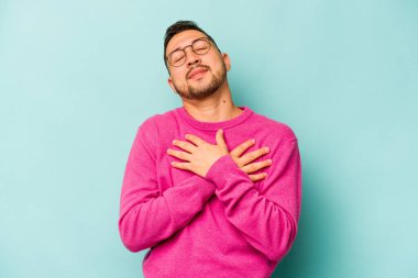 Mavi arka planda izole edilmiş genç İspanyol adam, avuçlarını göğsüne bastıran dostça bir ifadeye sahiptir. Aşk konsepti.