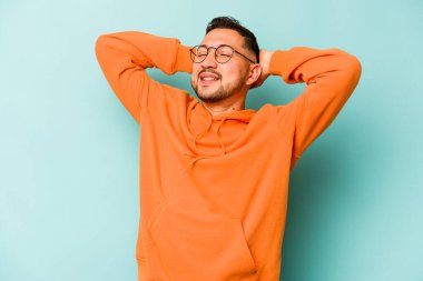 Genç İspanyol adam mavi arka planda izole edilmiş. Kendine güveniyor, elleri başının arkasında..