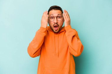 Genç İspanyol adam mavi arka planda izole edilmiş. Yüzünün yarısını avuç içiyle kaplayarak eğleniyor..