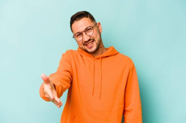 Mavi arka planda izole edilmiş genç İspanyol adam selamlama jesti olarak kameraya uzanıyor..
