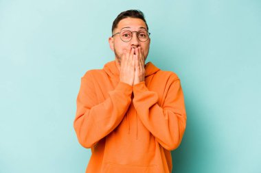 Mavi arka planda izole edilmiş genç İspanyol adam elleriyle ağzını kapatırken şok oldu..