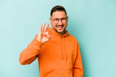 Genç İspanyol adam mavi arka planda izole edilmiş neşeli ve kendine güvenen iyi bir hareket sergiliyor..