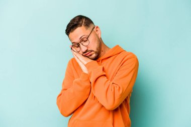 Mavi arka planda izole edilmiş genç İspanyol adam esniyor. Ağzı eliyle kapatan yorgun bir hareket sergiliyor..