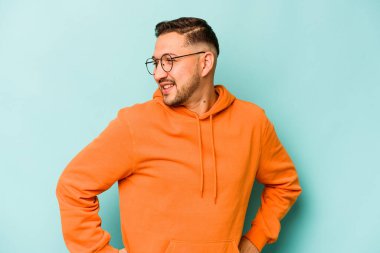 Latin kökenli genç adam mavi arka planda izole edilmiş. Kendine güveniyor. Kalçaları tutuyor..