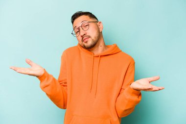 Genç İspanyol bir adam mavi arka planda izole edilmiş. Kafası karışmış ve omuz silkiyor. Kopya alanını tutmak için..