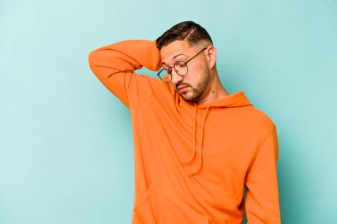 Genç İspanyol adam mavi arka planda izole edilmiş. Çok yorgun ve uykulu. Elini başının üstünde tutuyor..