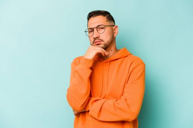 Genç İspanyol bir adam mavi arka planda izole edilmiş şüpheli, belirsiz, seni muayene ediyor..