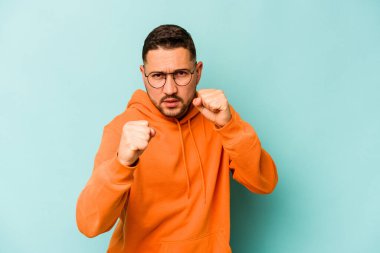 Mavi arka planda izole edilmiş genç İspanyol adam kameraya yumruk gösteriyor, agresif yüz ifadesi..