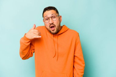 Genç İspanyol bir adam mavi arka planda izole edilmiş bir şekilde bir şeye gülüyor, elleriyle ağzını kapatıyor..