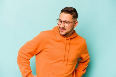 Mavi arka planda izole edilmiş genç İspanyol adam mutlu bir şekilde gülüyor ve ellerini karnının üstünde tutarak eğleniyor..