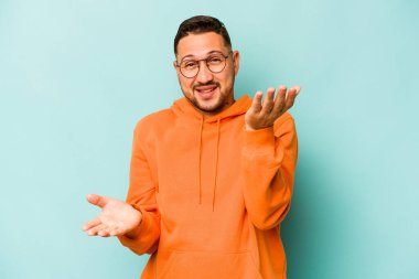 Mavi arka planda izole edilmiş genç İspanyol adam kollarıyla ölçeği yapıyor, mutlu ve kendinden emin hissediyor..