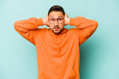 Genç İspanyol bir adam mavi arka planda izole edilmiş kulaklarını elleriyle kapatıyor çok fazla ses duymamaya çalışıyor..