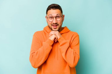 Latin kökenli genç adam mavi arka planda izole edilmiş şans için dua ediyor, şaşırmış ve ağzı açık bir şekilde önüne bakıyor..