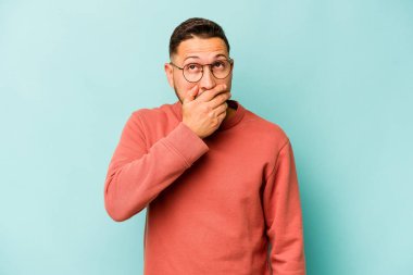 Genç İspanyol bir adam mavi arka planda izole edilmiş, ağzı kapalı ve elleri endişeli görünüyor..