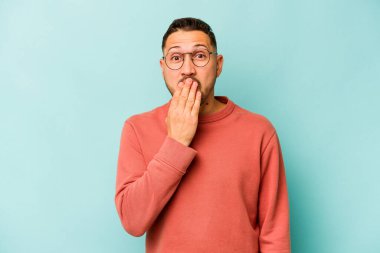 Mavi arka planda izole edilmiş genç İspanyol adam şok olmuş, ağzı ellerle kaplanmış, yeni bir şey keşfetmek için sabırsızlanıyor..