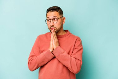 Genç İspanyol adam mavi arka planda izole edilmiş dua ediyor, bağlılık gösteriyor, dindar biri ilahi ilhamı arıyor..