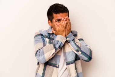 Beyaz arka planda izole edilmiş genç İspanyol adam parmaklarının arasından korkmuş ve gergin bir şekilde göz kırpıyor..