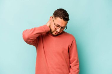 Genç İspanyol bir adam mavi arka planda izole edilmiş. Stres yüzünden boynu ağrıyor. Masaj yapıyor ve elle dokunuyor..